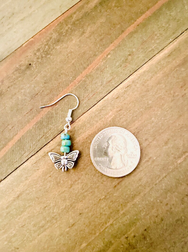 May include: Silver butterfly dangle earrings with turquoise blue beads. The earrings are shown next to a US quarter for size reference.