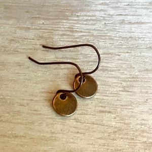 May include: A pair of small, round, bronze-colored metal disc earrings suspended from dark, curved wire hooks. Each circular charm features a smooth, slightly weathered finish and is attached to the hook through a small hole at the top edge.