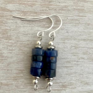May include: A pair of dangle earrings featuring stacked cylindrical blue lapis lazuli beads accented with small silver-toned metal spacers and beads. The earrings are suspended from simple silver-toned metal ear wires for a classic drop design.