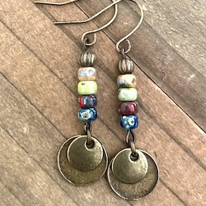 May include: A pair of handcrafted earrings featuring a cascade of colorful beads in shades of yellow, green, red, and blue. The beads are strung together and topped with a decorative metal bead and a pair of antique brass-colored coin charms.