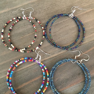 May include: Four pairs of beaded hoop earrings. The earrings are made with small, colorful beads and have silver hooks. The earrings are arranged in a row on a wooden surface.