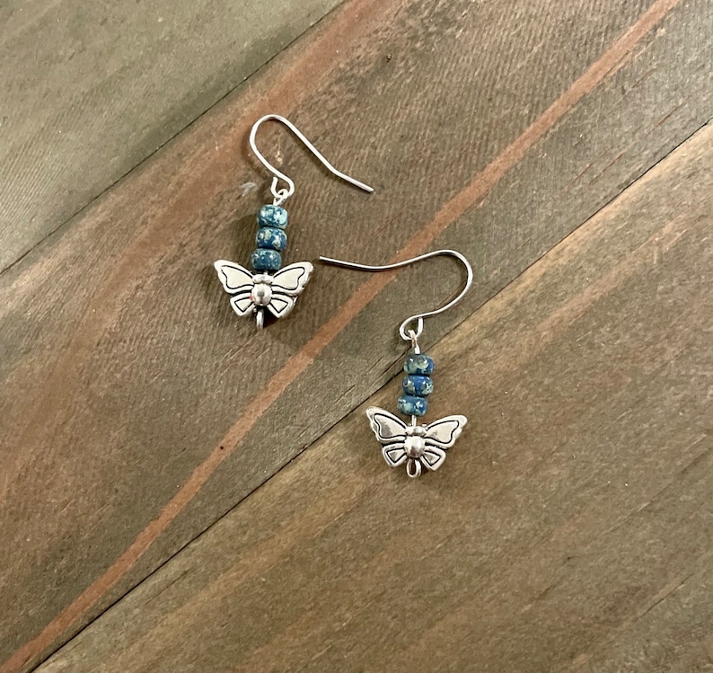 May include: A pair of silver butterfly earrings with blue and white speckled beads hanging from the bottom.