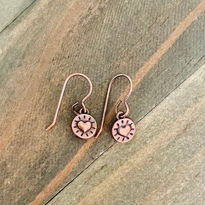 May include: A pair of copper earrings with a small heart design on a circular charm. The earrings have a simple hook closure.