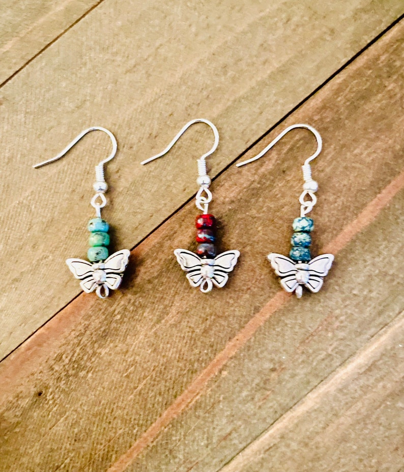 May include: Three pairs of silver butterfly earrings with colorful stone beads. The earrings have a silver butterfly charm at the bottom and a silver hook closure.