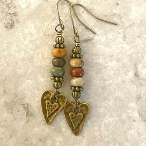 May include: A pair of dangle earrings with a heart charm. The earrings feature brown, green, and beige stone beads.