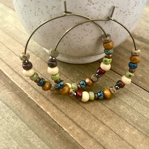 May include: A pair of hoop earrings with a variety of colorful beads. The beads include shades of brown, green, red, and blue, interspersed with cream-colored beads. The hoops are made of a gold-colored metal.