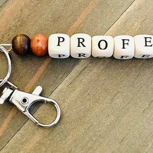 May include: A silver keychain with a metal ring and a clasp. The keychain has a wooden bead charm with the word "PROFE" spelled out in black letters.