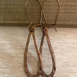 May include: A pair of copper-colored teardrop earrings. The earrings have a hammered texture and a long, curved hook. The teardrop shape is elongated and slightly irregular, giving them a handcrafted appearance. The earrings are displayed against a light wood background.