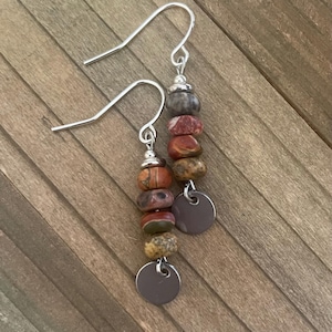 May include: A pair of dangle earrings featuring silver-toned hooks and small metal spacers. Each earring is composed of a vertical stack of four multi-colored, earth-toned stone beads in shades of brown, red, and gray, finished with a flat metal disc.