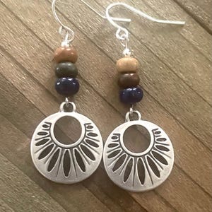 May include: A pair of silver-toned earrings with a circular, sunburst-like design. Each earring features a series of colorful beads in shades of brown, green, and blue. The earrings hang from silver-colored ear wires, creating a bohemian aesthetic.