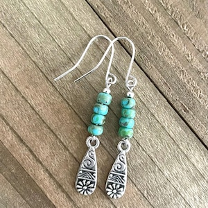 May include: A pair of turquoise and silver dangle earrings. The earrings feature a teardrop-shaped pendant with a silver design and a row of turquoise beads.