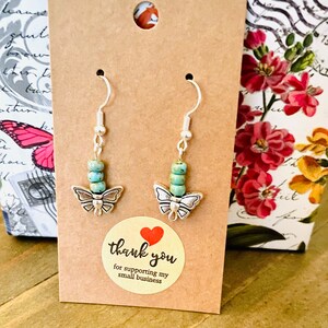 May include: A pair of silver butterfly earrings with turquoise beads. The earrings are displayed on a brown card with a thank you sticker.
