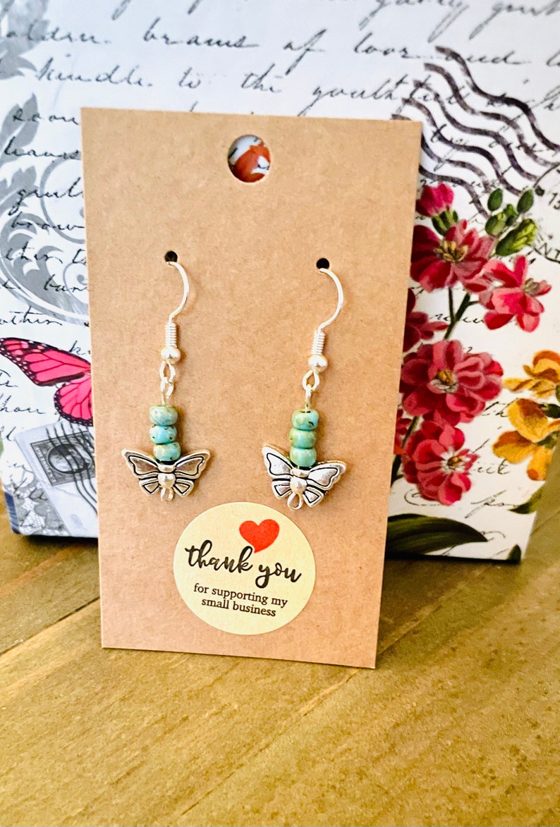 May include: A pair of silver butterfly earrings with turquoise beads. The earrings are displayed on a brown card with a thank you sticker.