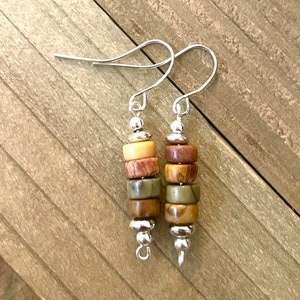 May include: A pair of dangle earrings featuring stacked, cylindrical beads in shades of beige, brown, and green. The beads are separated by small, silver-colored metal accents and suspended from silver-colored ear wires.