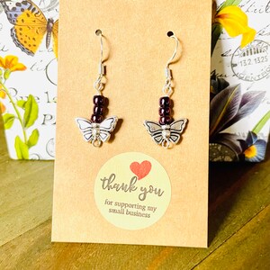 May include: A pair of silver butterfly earrings with dark purple beads hanging from the hooks. The earrings are displayed on a brown card with the text "thank you for supporting my small business".
