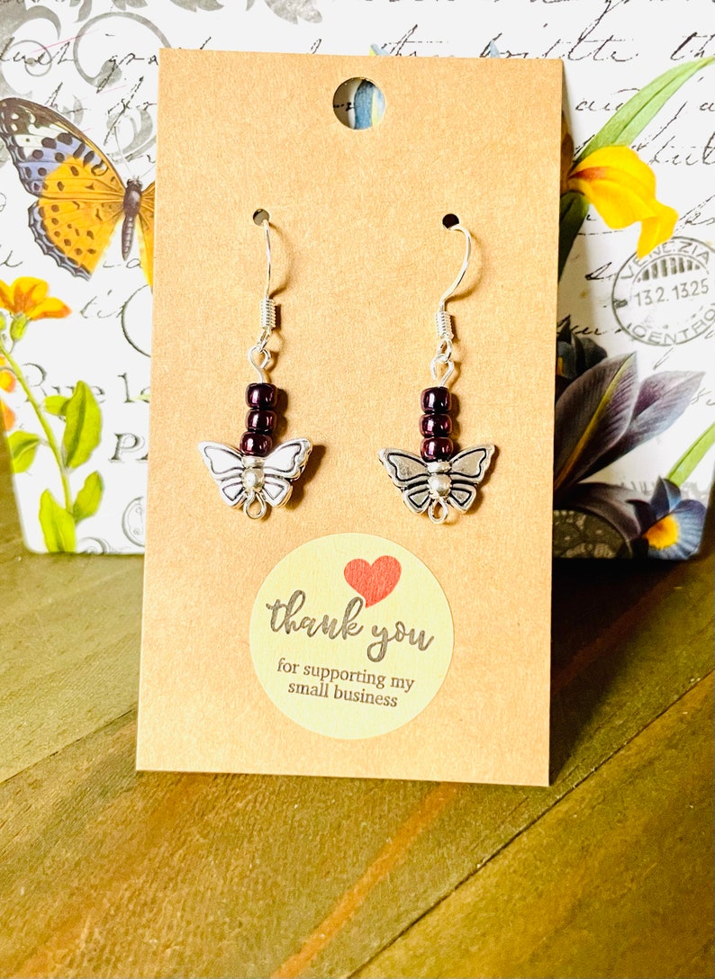 May include: A pair of silver butterfly earrings with dark purple beads hanging from the hooks. The earrings are displayed on a brown card with the text "thank you for supporting my small business".