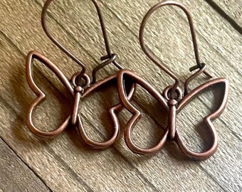 May include: A pair of copper-colored butterfly earrings. Each earring features a butterfly outline with a hook for pierced ears. The earrings are displayed on a wooden surface, showcasing their delicate design and warm metallic hue.