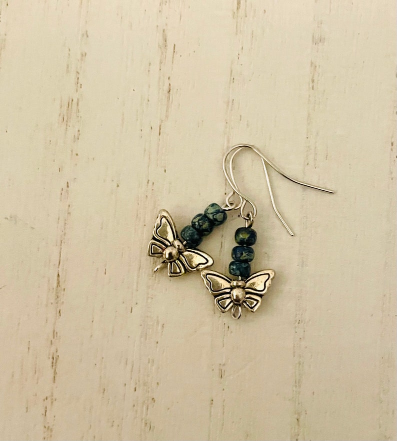 May include: A pair of silver butterfly earrings with blue and green speckled beads hanging from the bottom.