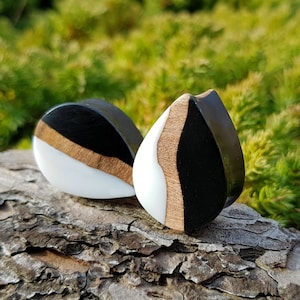 May include: Two teardrop-shaped wooden ear gauges with black, white and brown resin. The gauges are on a wooden surface.