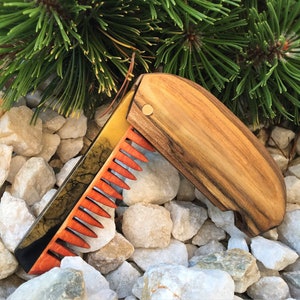 Könnte beinhalten: Ein Kamm aus Holz mit einer schwarzen und goldenen Einlage und orangenen Zähnen. Der Kamm ist zusammengeklappt und liegt auf einem Bett aus weißen Steinen.