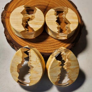 May include: Four wooden and resin ear gauges with a clear resin inlay. The gauges are in the shape of a circle and an oval. The wood is a light brown color with a natural wood grain pattern. The resin is clear and has a slight shimmer.