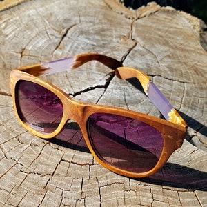 Puede incluir: Gafas de sol de madera con lentes tintados de color morado y un marco de madera marr&oacute;n claro. Las gafas de sol est&aacute;n sobre una superficie de madera.