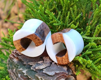 May include: Two white and brown wooden ear gauges. The gauges are round and have a hole in the center. The gauges are on a brown and green surface.