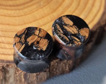 May include: A pair of circular ear plugs featuring a combination of natural brown wood grain and glossy black resin. The design displays an organic, fractured pattern where the dark resin fills the spaces between the textured wood segments.