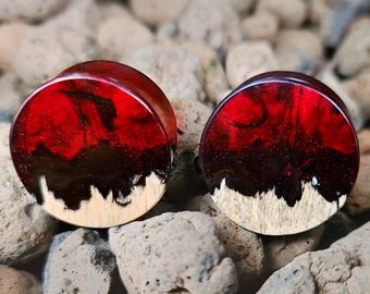 May include: Pair of red and black resin ear plugs with a wood base. The plugs have a marbled effect with a dark red and black design. The base is a light wood color with a jagged edge.