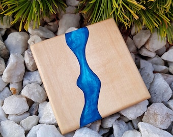 May include: A square wooden coaster with a blue resin river design. The coaster is made of light wood and has a natural finish.