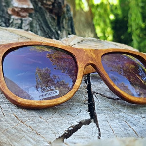 Puede incluir: Gafas de sol de madera marr&oacute;n con un patr&oacute;n de veta de madera marr&oacute;n claro. Las lentes est&aacute;n te&ntilde;idas de morado y reflejan el cielo y los &aacute;rboles. Las gafas de sol tienen una etiqueta blanca que dice "Udeo UV Protection".