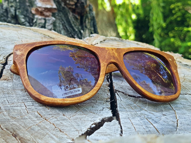 Puede incluir: Gafas de sol de madera marr&oacute;n con un patr&oacute;n de veta de madera marr&oacute;n claro. Las lentes est&aacute;n te&ntilde;idas de morado y reflejan el cielo y los &aacute;rboles. Las gafas de sol tienen una etiqueta blanca que dice "Udeo UV Protection".