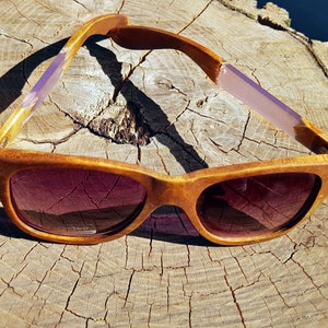 Puede incluir: Gafas de sol de madera con monturas marrones y detalles morados. Las gafas de sol est&aacute;n sobre una superficie de madera.