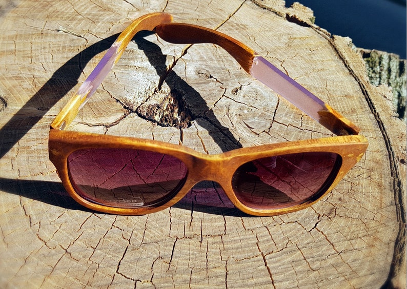 Puede incluir: Gafas de sol de madera con monturas marrones y detalles morados. Las gafas de sol est&aacute;n sobre una superficie de madera.