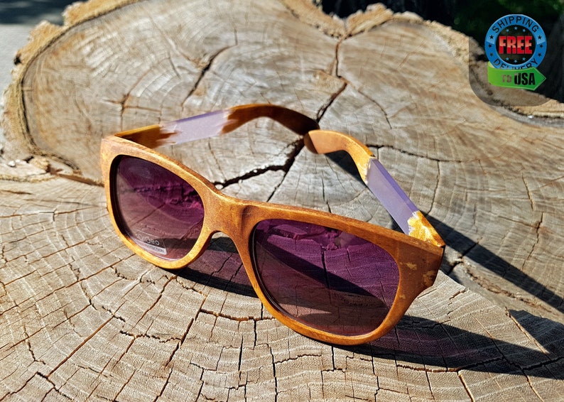 Puede incluir: Gafas de sol de madera con lentes tintados de color morado y un marco de madera marr&oacute;n claro. Las gafas de sol est&aacute;n sobre una superficie de madera.