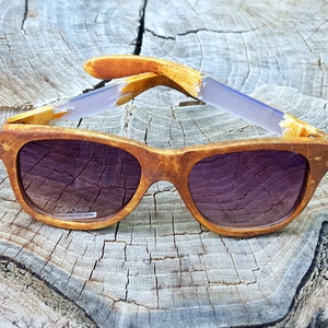 Puede incluir: Gafas de sol de madera con lentes marrones y un marco de madera marr&oacute;n claro. Las gafas tienen un acento morado claro en las patillas. El texto "ecoo" y "UV400 PROTECTION" est&aacute; impreso en la lente derecha.