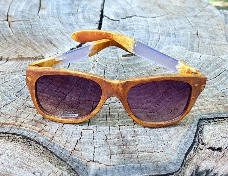 Puede incluir: Gafas de sol de madera con lentes marrones y un marco de madera marr&oacute;n claro. Las gafas tienen un acento morado claro en las patillas. El texto "ecoo" y "UV400 PROTECTION" est&aacute; impreso en la lente derecha.
