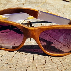 Puede incluir: Un par de gafas de sol de madera con lentes tintados de color morado. Las gafas est&aacute;n hechas de madera marr&oacute;n claro y tienen un acabado natural. Las patillas de las gafas est&aacute;n hechas de una madera m&aacute;s clara y tienen una franja blanca.