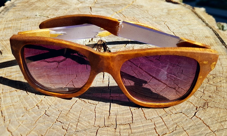 Puede incluir: Un par de gafas de sol de madera con lentes tintados de color morado. Las gafas est&aacute;n hechas de madera marr&oacute;n claro y tienen un acabado natural. Las patillas de las gafas est&aacute;n hechas de una madera m&aacute;s clara y tienen una franja blanca.