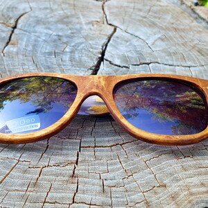Puede incluir: Gafas de sol de madera con monturas marrones y lentes oscuras. Las lentes reflejan el cielo y los &aacute;rboles. Las gafas de sol tienen una etiqueta "Urideo Protection" en la lente derecha.