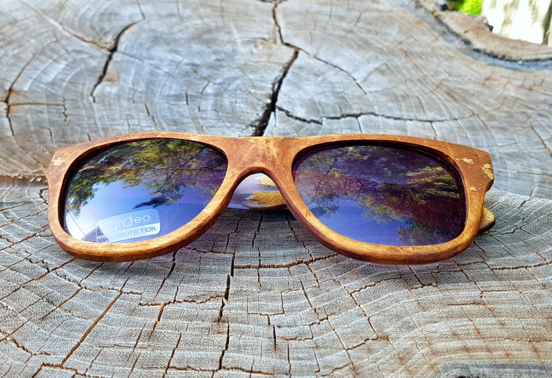 Puede incluir: Gafas de sol de madera con monturas marrones y lentes oscuras. Las lentes reflejan el cielo y los &aacute;rboles. Las gafas de sol tienen una etiqueta "Urideo Protection" en la lente derecha.
