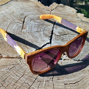 Puede incluir: Un par de gafas de sol de madera con detalles en morado y dorado. Las gafas tienen una montura marr&oacute;n y lentes rosas. Las gafas est&aacute;n sobre una superficie de madera.