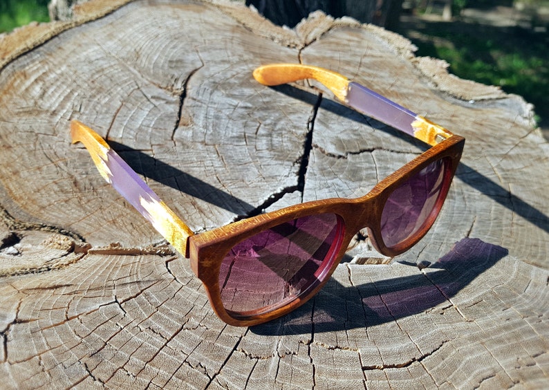 Puede incluir: Un par de gafas de sol de madera con detalles en morado y dorado. Las gafas tienen una montura marr&oacute;n y lentes rosas. Las gafas est&aacute;n sobre una superficie de madera.