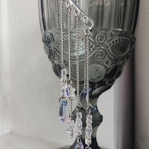 May include: A silver ear cuff with a delicate wing design and dangling chains adorned with clear and iridescent beads. The ear cuff is displayed next to a gray glass goblet, creating a whimsical and ethereal aesthetic.