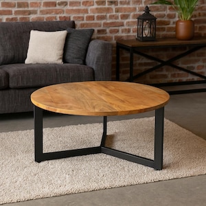 Op de afbeelding: Een ronde salontafel met een lichtbruin houten blad en zwarte metalen poten. De tafel staat op een beige tapijt. Het ontwerp is modern en minimalistisch, geschikt voor een woonkamer.