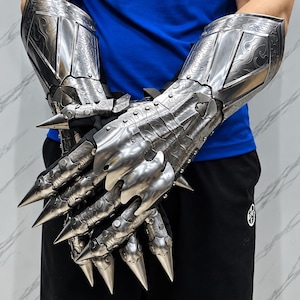 Könnte beinhalten: Ein Paar bewegliche Metallhandschuhe mit komplizierten Schnörkelgravuren und langen, spitzen, krallenartigen Fingerverlängerungen. Die silberfarbene Rüstung bedeckt die Hände und Unterarme und verfügt über segmentierte Platten für Beweglichkeit.