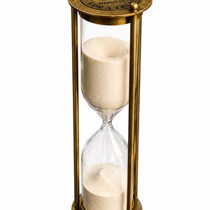 May include: A 60-minute hourglass with a metallic brass frame and clear glass bulbs containing beige sand. The top circular brass plate features the engraved text SAND TIMER 60 MINUTES FOR A TRUST, with small decorative spheres at the edges.