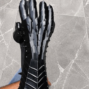 K&ouml;nnte beinhalten: Ein schwarzer, beweglicher Panzerhandschuh mit einem geschichteten, schuppenartigen Design, das Unterarm, Handgelenk und Finger bedeckt. Die R&uuml;stung besteht aus mehreren &uuml;berlappenden Platten, die mit kleinen Nieten befestigt sind und eine segmentierte Struktur bilden.