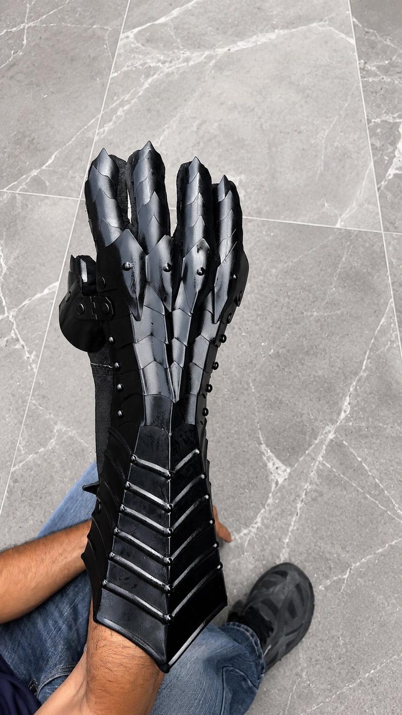 K&ouml;nnte beinhalten: Ein schwarzer, beweglicher Panzerhandschuh mit einem geschichteten, schuppenartigen Design, das Unterarm, Handgelenk und Finger bedeckt. Die R&uuml;stung besteht aus mehreren &uuml;berlappenden Platten, die mit kleinen Nieten befestigt sind und eine segmentierte Struktur bilden.