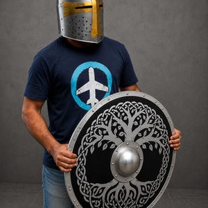 Op de afbeelding: Een zilveren ridderhelm met een gouden kruisaccent en een rond zwart schild met een zilveren levensboomontwerp en ingewikkelde knooppatronen. Het schild heeft een metalen rand en een centrale koepel, vastgehouden naast een donkerblauw t-shirt.